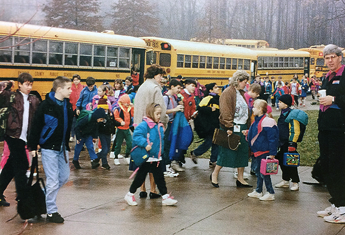 Photograph of students arriving at the start of the school day at Forestville. School buses are parked along the sidewalk in front of the building, and children are walking toward the front doors of the school.
